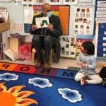 A classroom of preschool students listen to an adult read a book